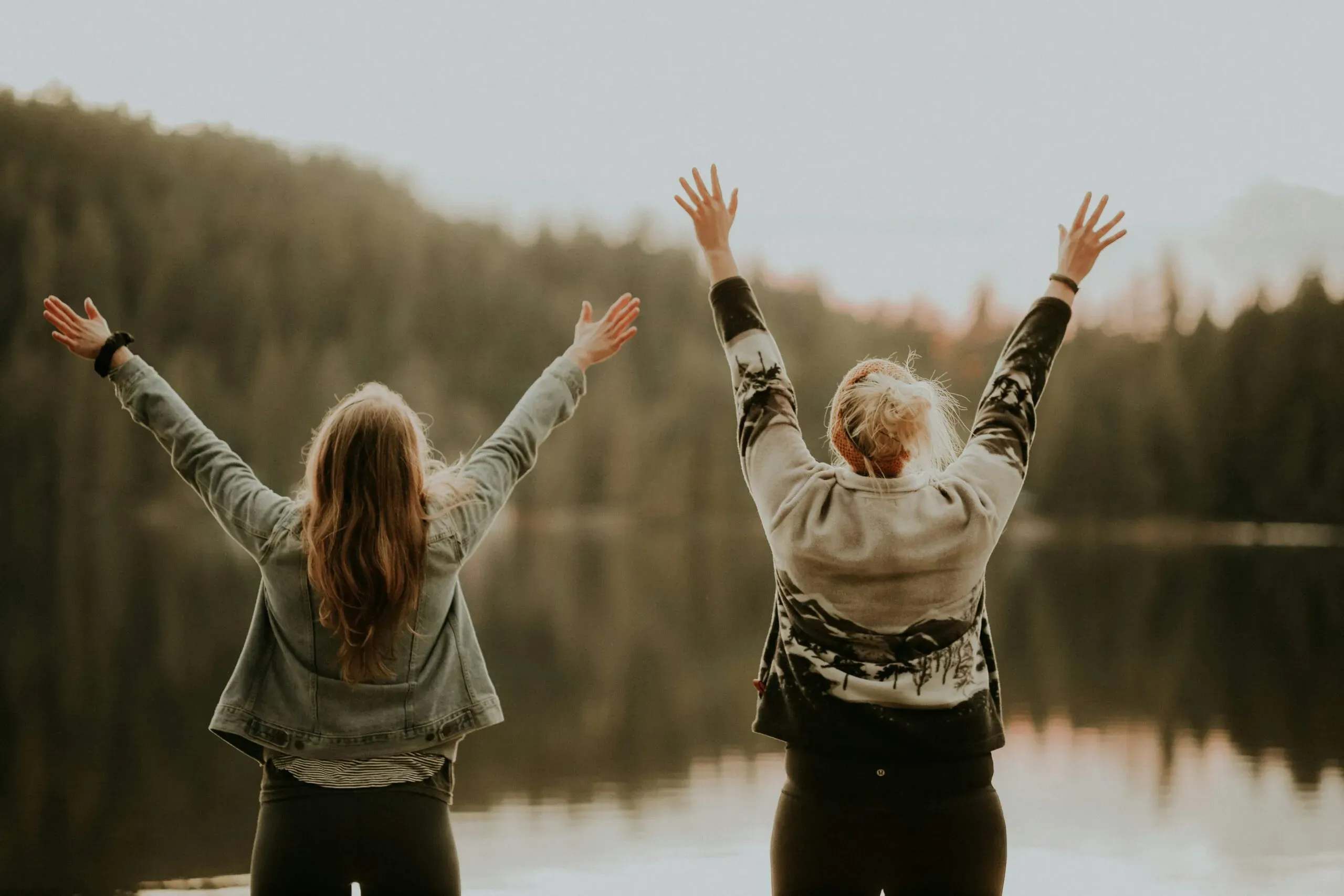 Deux femmes de dos levant les bras en l'air face à un lac calme et des montagnes boisées.